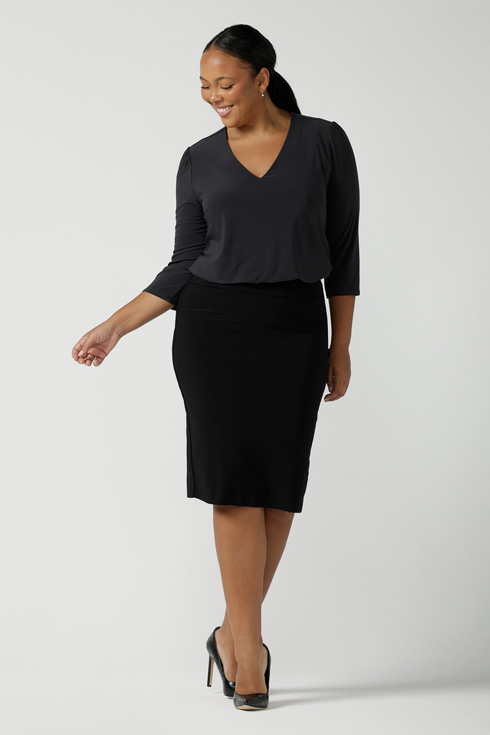 Size 16 woman wears the Vida top in Charcoal, a grey work top for women with a V-neckline. Comfortable corporate workwear for women. Made in Australia for women. Size 8 - 24. Styled back with a black slim fit Andi skirt and black pumps.