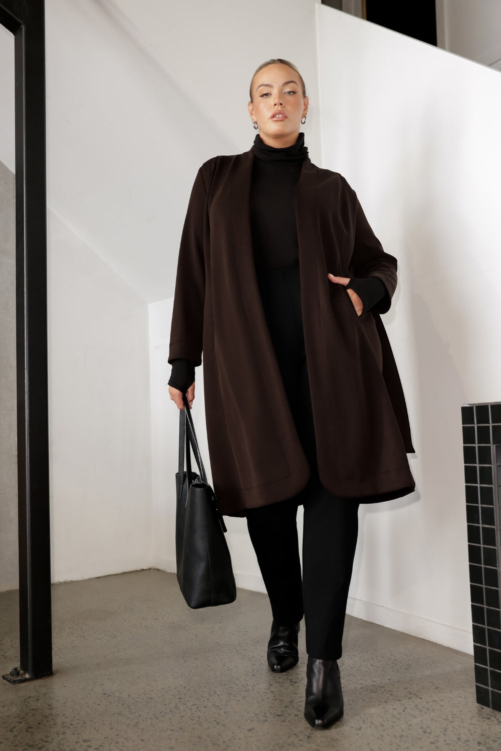 The Sorel Trench Coat is expertly tailored with a modern, sophisticated silhouette that elevates any wardrobe. Size 18 woman wears winter coat crafted from a luxurious winter-weight Modal fabric over long length black pants, black turtle neck, and high heel boots for an elevated workwear look. 