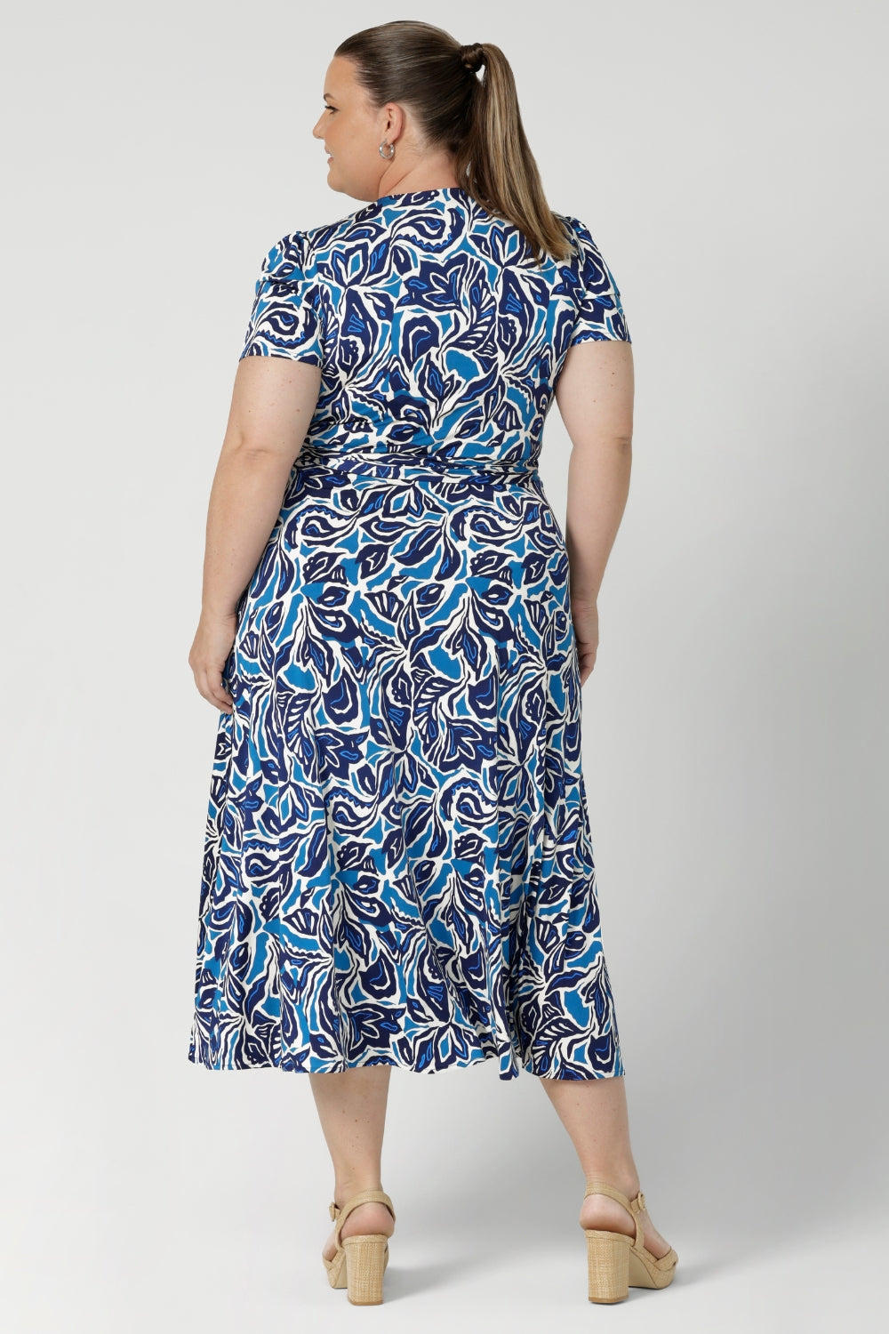 Plus size model wears short sleeve dress, designed with delicate tucks on the shoulder, midi length skirt & full wrap design. From workday sophistication to event-ready weekend comfort. This dress is built on a slinky jersey base, offering a soft, stretchable fit that moves with everybody to ensure comfort and style. Available in sizes 8-24.