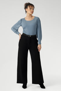 Size 10 Woman wears black wide leg work pants with a blue blouse tucked in and black boots. These black pants feature wide legs, belt loops, high waist and tailored details for excellent shaping. Available in sizes 8-24.