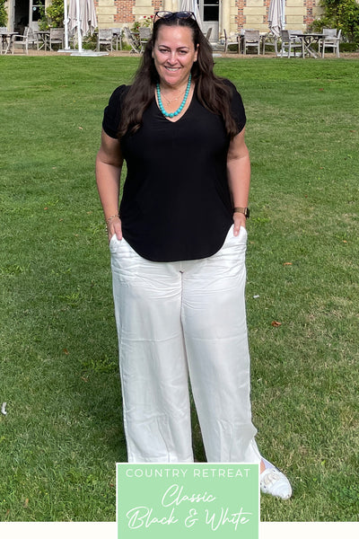 Showing casual travel wear for a holiday in France, a plus size, curvy woman wears a V-neck, black short sleeve top in bamboo jersey with white wide leg pants in sustainable tencel fabric, both made in Australia by women's clothing brand Leina & Fleur.