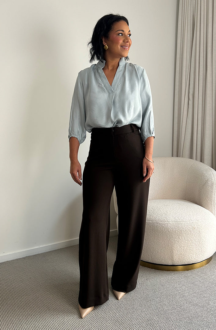 Showing a work wear outfit for professional women, a size 10 woman wears an ice blue pull-on shirt with 3/4 sleeves and brown wide leg pants. Both blouse and pants for women are made in Australia in sizes 8-24 by women's clothing brand, Leina + Fleur. 