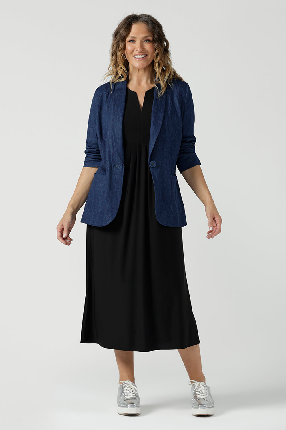 A size 10 woman wears the Vintage Denim Xavier Blazer. A button front style blazer with pockets. Made in Australia for women size 8-24. Styled back with a black jersey dress.