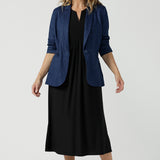 A size 10 woman wears the Vintage Denim Xavier Blazer. A button front style blazer with pockets. Made in Australia for women size 8-24. Styled back with a black jersey dress.