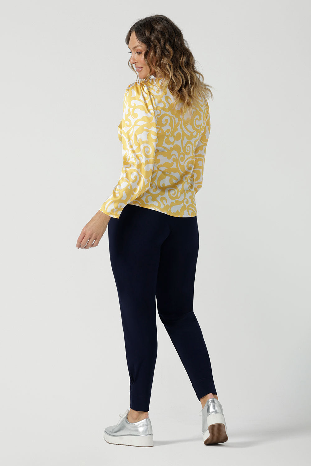 Back view of a size 10 woman wears a Zara Top in Yellow Swirl silk. A long sleeve blouse with fashion sleeve and made in 100% silk. Made in Australia for women size 8-24. Styled back with the Indi Tall Pant in Navy.