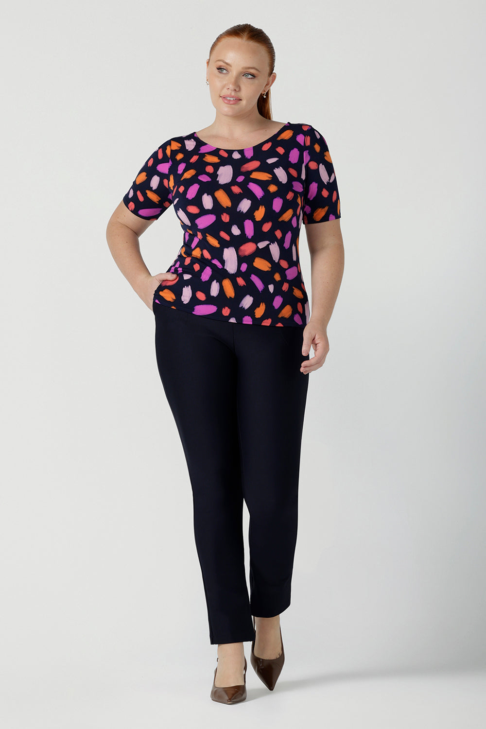 Size 12 woman wears the Ziggy top in Palette. A boat neckline style with short sleeves and shoulder tucks. A great work to weekend top for women. Palette print features a brush stroke print of fuchsia, lilac, red and orange . Made in Australia for women size 8 to 24.Styled back with Brooklyn pants in Navy. 