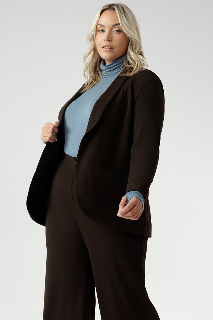 curvy woman wears a plus size brown blazer over a sky blue turtle neck top and brown wide leg pants.