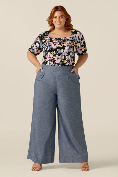 A size 18 woman wears a short-sleeve, square scoop neck top in a floral print jersey. The top is worn tucked in to high-waisted wide-leg pants in denim-like chambray fabric. Both are made in Australia for women between sizes 8 and sizes 24.