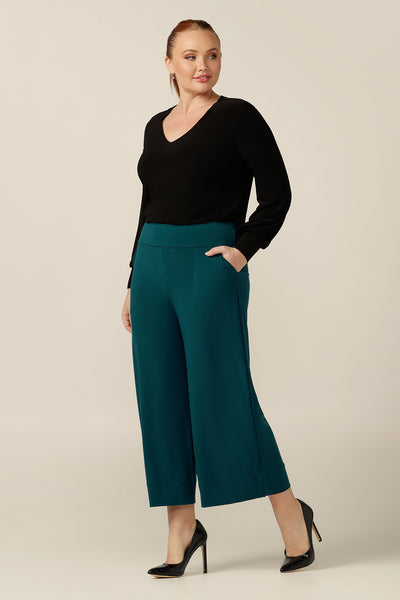 a size 12 curvy woman wears a wide-leg pull-on pant in Petrol green jersey. A cropped length, culotte trouser with extra wide legs, this stretch pant is worn with a long sleeve black top and heels for a corporate wear look. 
