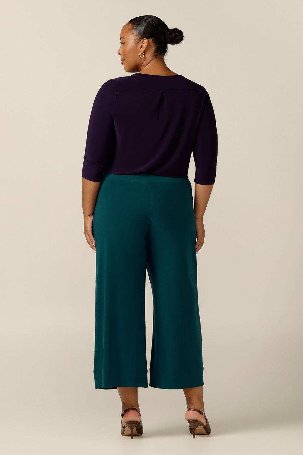 a plus size, size 18 curvy woman wears a wide-leg pull-on pant in Petrol green jersey. A cropped length, culotte trouser with extra wide legs, this stretch pant is worn with a long sleeve purple top and heels for a work wear wear look. 