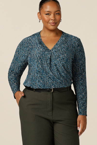 A size 18, fuller figure woman wears a long sleeve, V-neck jersey top in blue, black and white printed jersey jacquard. Tucked in to olive green tailored work pants, this long sleeve top for women works for work and casual wear.