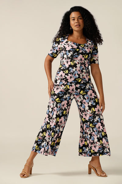 A sizes 12 woman wears a short-sleeve printed jersey top with matching wide-leg printed pants to create a faux jumpsuit look. The top has a squared scoop neckline and supported bust. The trousers are a pull-on jersey pant cropped in length. Both the women's top and pants feature a floral print with a black base and pastel flowers. Both are available to shop online from sizes 8 to size 16 and plus sizes 18 to size 24 at Leina and Fleur Australia.
