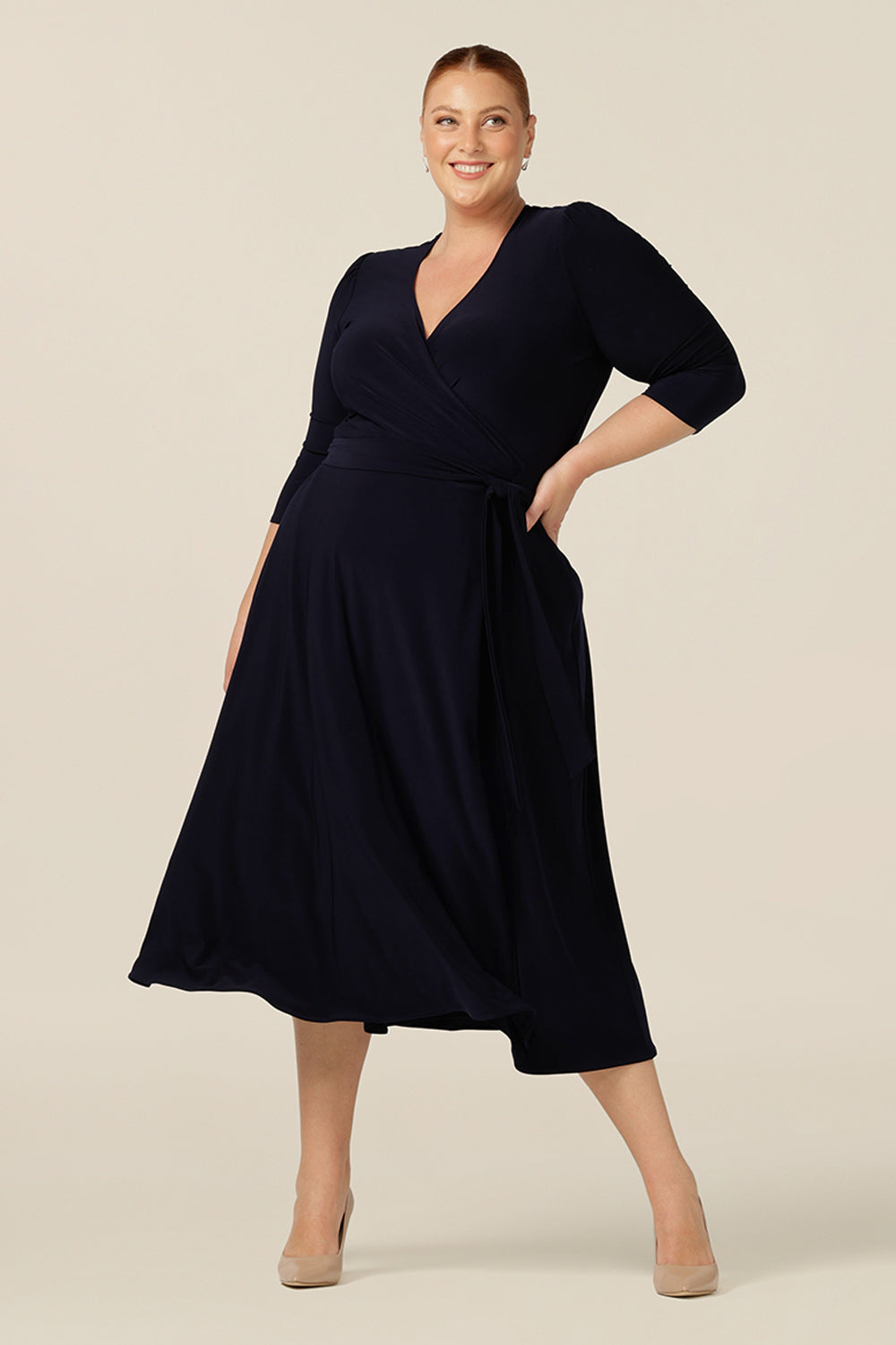 A plus size, fuller figure woman wears a 3/4 sleeve, jersey wrap dress in navy blue. A full, below-the-knee-length wrap dress, the Fiona Dress wears well as an elegant workwear dress as well as for occasion and event wear. 