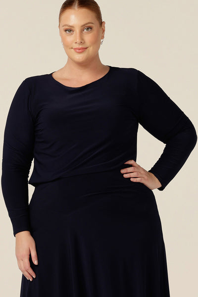 A plus size, size 18 woman wears a boat neck top in navy blue jersey. Featuring long sleeves, this basic women's top is worn with a navy skirt as a faux dress look.