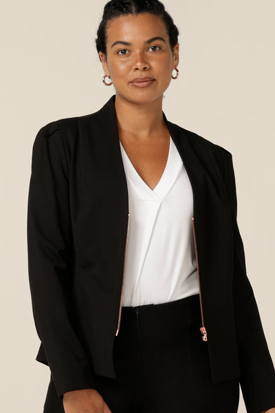 A size 12, curvy woman wears a corporate black jacket with white bamboo jersey top. A soft tailored jacket with rose gold zip fastening, long sleeves and a collarless neckline, it is worn with black tailored trousers. 