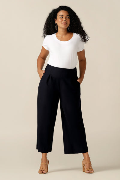A size 12 curvy woman wears wide-leg, cropped length pants in Navy. Worn with a white top in bamboo jersey, the pants are styled as modern work wear trousers. With front pleats, side pockets and in stretch jersey, they are cut as a comfortable fit pant for all-day corporate wear. 
