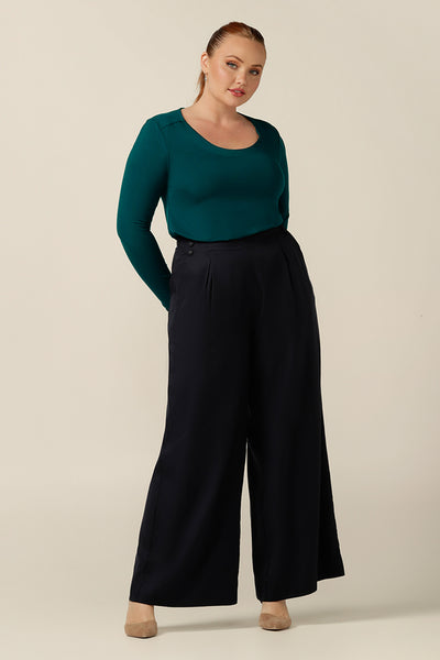 a size 12, curvy woman wears a round neck, long sleeve, fitted top in green bamboo jersey. In natural fibres, the bamboo jersey top is worn with wide leg navy blue pants - both top and trousers are lightweight, breathable and comfortable for corporate or casual wear.
