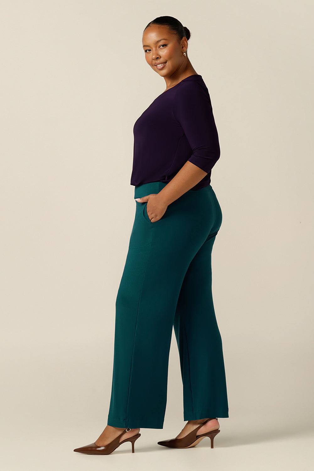a size 18 curvy woman wears a wide-leg, pull-on pant in Petrol green jersey. A full length length, straight cut wide trouser, this stretch pant is worn with a long sleeve purple top and heels for a corporate wear look. 