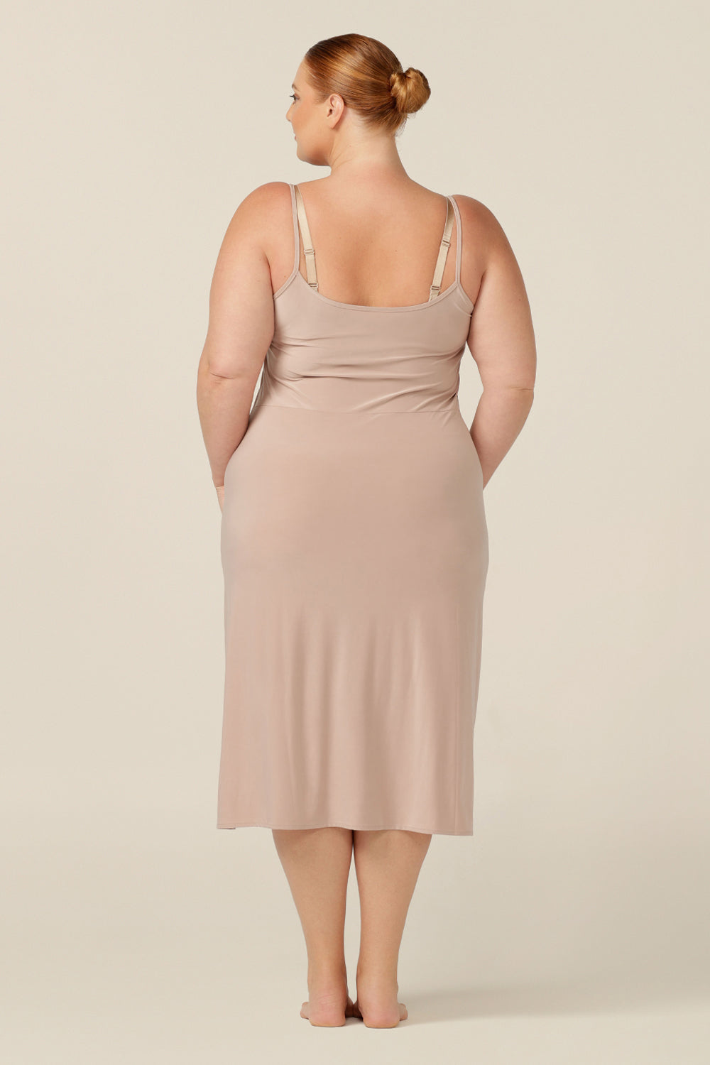 A fuller figure woman wears a flesh-coloured, midi-length, reversible slip in slinky jersey. Worn with a V neck to the front, and scoop neck to the back. With a skirt length finishing below the knee, this midi slip gives a smooth layer under dresses and skirts.