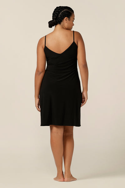 A size 12 woman wears a short, above-the-knee length, reversible slip in black, slinky jersey fabric. The slip has thin straps and is worn with a scoop neck to the front, V neckline to the back. plus size strap dress