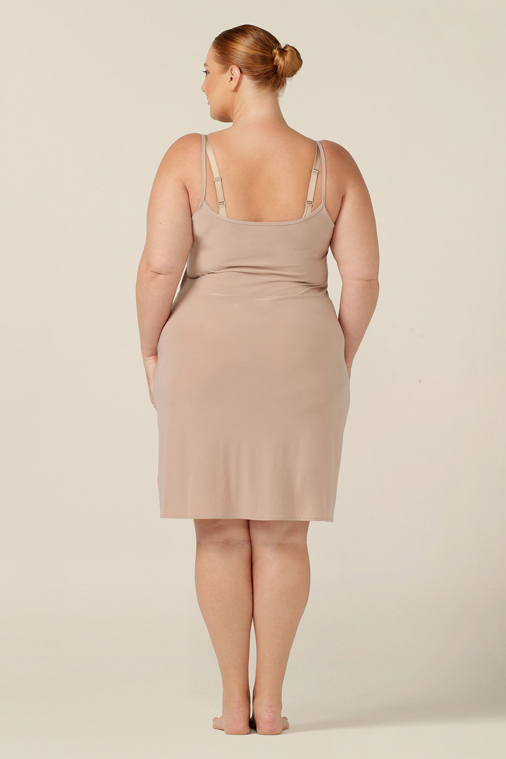 back view of a fuller figure, size 18 woman wearing a neutral, mini-length slip. A reversible nude-coloured slip, this undergarment can have a V-neck or a scoop neck to give a smooth layer under dresses and skirts. Australian women's fashion that is perfect for wearing around the house as loungewear.