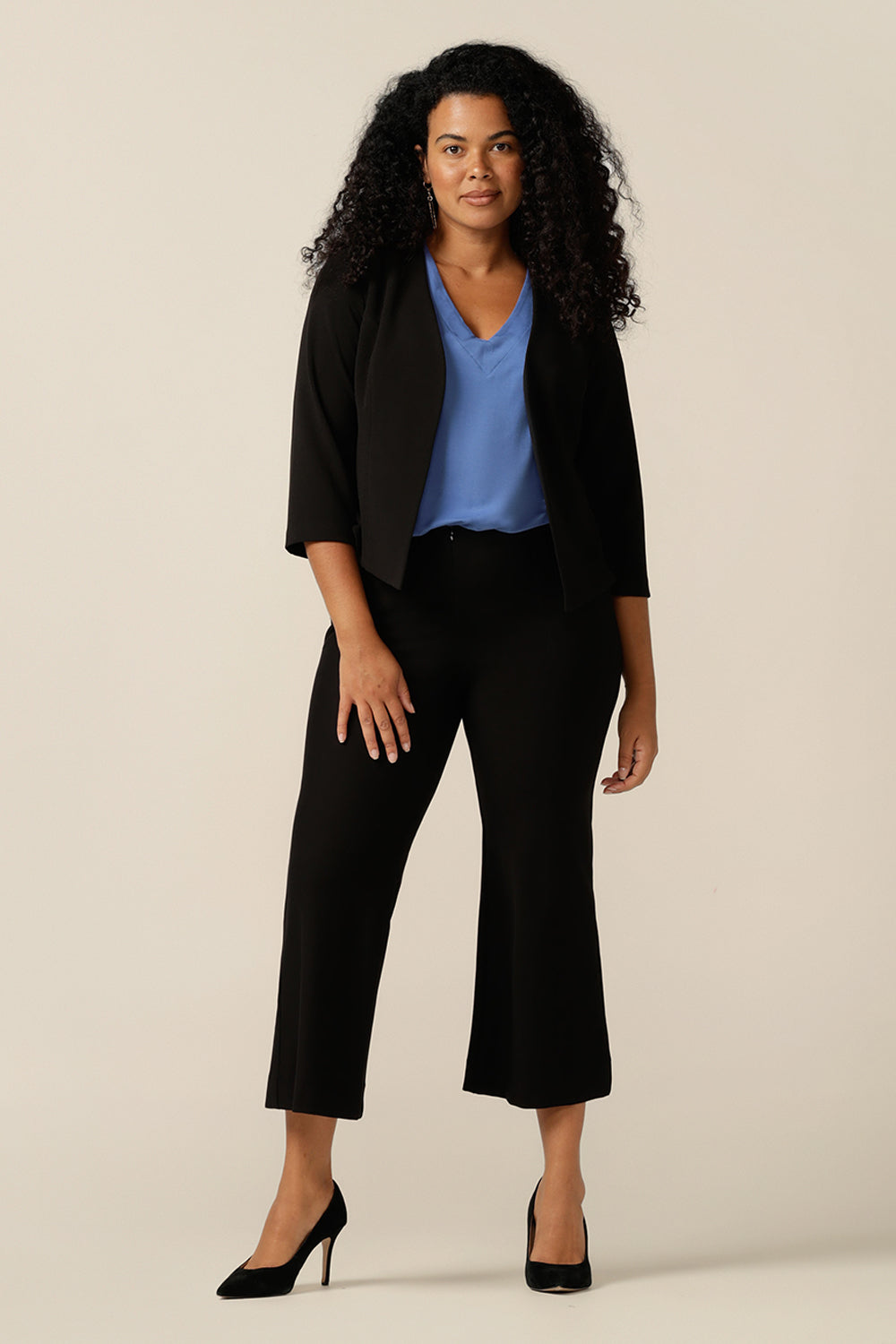 A size 12 woman wears a collarless, open-front black jacket with a blue bamboo jersey top and cropped, tailored black trousers for work. Made in Australia in stretch jersey, this corporate jacket is made from stretch jersey for a comfortable jacket fit. 