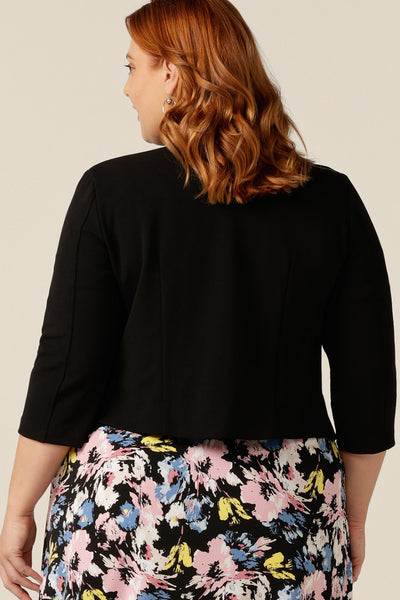 A plus size, size 18 woman wears a black workwear jacket over a floral print jersey dress. The Rainy Jacket is a collarless, open front jacket with cropped sleeves. Made in crepe jersey, this is a soft tailoring jacket with stretch for a comfortable twist on quality-made work wear suit jackets for women.  